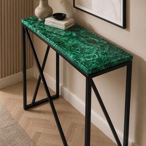 Puede incluir: Una mesa consola rectangular con una superficie superior de patrón de malaquita verde vibrante y un marco de metal negro con soportes laterales en forma de X. La mesa sostiene un jarrón beige con vegetación y dos libros con el título Live Beautifully.
