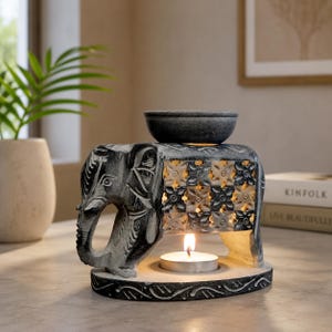 May include: A gray and black elephant-shaped oil burner featuring a carved floral pattern on the side and a circular bowl on top. A small tea light candle is lit underneath the elephant, casting a warm glow through the cut-out design.