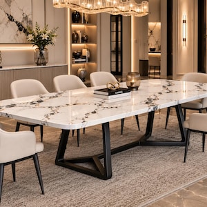 May include: A rectangular dining table with a white marble top featuring black veining and a black metal base, surrounded by six cream-coloured upholstered chairs with black legs. Two books and a gold decorative object sit on the table surface.