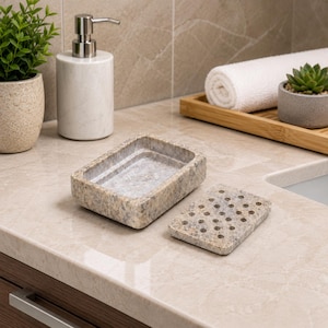 May include: A rectangular soap dish made of speckled beige and grey stone, featuring a separate removable lid with a grid of circular drainage holes. The dish has a smooth, polished finish and a natural, mottled appearance with earthy tones.