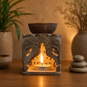 May include: A square, light brown stone oil burner featuring carved elephant motifs on the front. The structure has an arched opening containing a lit tea light candle, with a small, matching stone bowl resting on the top surface for oil.