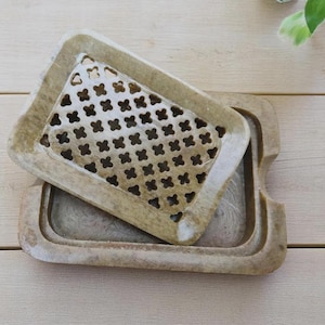 May include: A rectangular soap dish made of light brown stone with a smooth finish. The dish consists of two parts: a base tray with a recessed center and a removable top insert featuring a grid of small, cross-shaped cutouts for drainage.