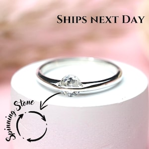Pode incluir: Um anel de prata com uma pedra preciosa clara e redonda. O anel é exibido em um suporte cilíndrico branco sobre um fundo rosa. O texto "SHIPS NEXT DAY" é visível na parte superior. As palavras "Spinning Stone" estão escritas no lado esquerdo.