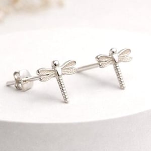 May include: A pair of silver-toned dragonfly stud earrings featuring detailed wing patterns and a segmented body design. Each earring includes a post back closure, showcasing a metallic finish with intricate textures on the wings and tail.