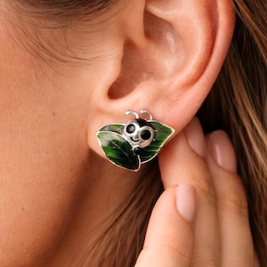 May include: Silver stud earrings with a ladybird design. The ladybird has a black body and a smiling face, set on a green leaf. These earrings are a charming accessory.