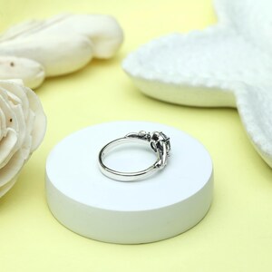 May include: A silver ring with an ornate design, featuring a dark gemstone at the center. The ring is set on a white circular platform against a soft yellow background. The ring's intricate details and the gemstone's color are highlighted.