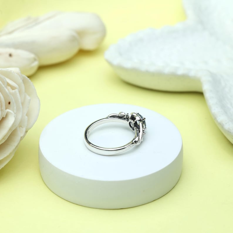 May include: A silver ring with an ornate design, featuring a dark gemstone at the center. The ring is set on a white circular platform against a soft yellow background. The ring's intricate details and the gemstone's color are highlighted.