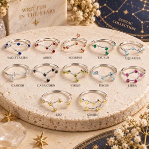 May include: A collection of twelve silver-toned rings, each featuring a unique constellation design with small coloured gemstones. The rings are labelled with zodiac signs including Sagittarius, Aries, Scorpio, Taurus, Aquarius, Cancer, Capricorn, Virgo, Pisces, Libra, Leo, and Gemini.