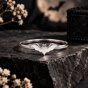 Puede incluir: Un anillo de tono plateado pulido que presenta un emblema central con forma de murciélago. La banda es delgada y circular, con el motivo del murciélago integrado en la parte superior, mostrando superficies lisas y brillantes.