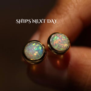 May include: Two round stud earrings featuring bezel-set opals with iridescent flashes of green, orange, and pink set in polished gold-tone metal. The text SHIPS NEXT DAY is visible in white lettering above the earrings.