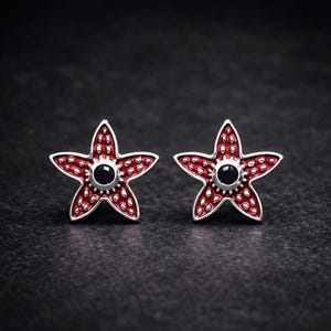 Puede incluir: Un par de pendientes de estrella de mar plateados con detalles de esmalte rojo y una piedra negra en el centro. Los pendientes tienen forma de estrella de cinco puntas con pequeños puntos en relieve alrededor de los bordes. Fondo oscuro.