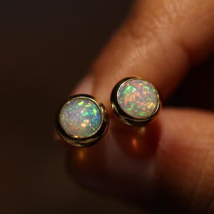 May include: Two gold stud earrings, each with a round opal gemstone. The opals have a play of colour, with green, red, and blue hues, set in a gold bezel. The earrings are displayed against a blurred background.