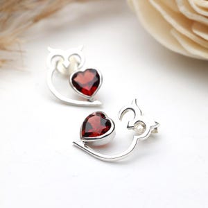 May include: A pair of silver stud earrings. Each earring has a heart-shaped, deep red gemstone set in a silver frame. The design incorporates a stylized owl shape above the heart, creating a unique and elegant appearance.