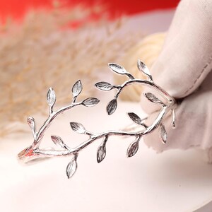 May include: Silver-toned bracelet featuring a delicate leaf design. The bracelet has a curved shape, with branches extending across the top and adorned with detailed leaves. The metal has a polished finish, reflecting light and adding a touch of elegance.