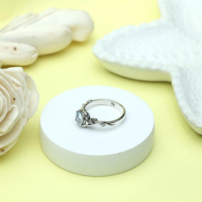 May include: A silver ring with a light blue gemstone center. The ring features a floral design with delicate leaves and small accents. The letter "N" is engraved on the band. The ring is displayed on a white circular platform against a yellow background.