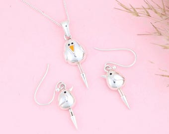 Puede incluir: Un conjunto de joyas plateadas con forma de pájaro, que incluye un collar con colgante y dos pendientes. El pájaro tiene un cuerpo blanco, un pequeño pico naranja y una cola puntiaguda. Las joyas se exhiben sobre una superficie rosa.