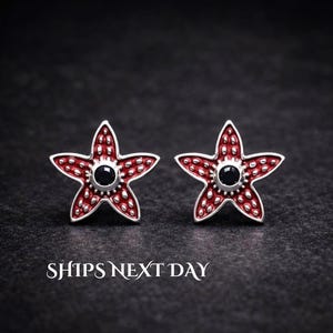 May include: A pair of star-shaped stud earrings featuring a red textured surface with silver-toned raised dots and a central black circular stone. The text SHIPS NEXT DAY is printed in white at the bottom of the image.