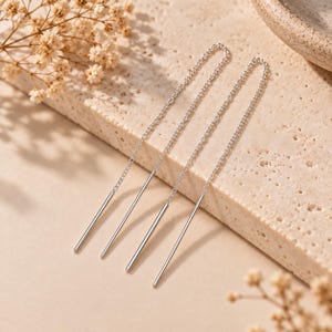 May include: A pair of silver threader earrings featuring a delicate fine chain connected to a slender, straight metal bar at each end. The earrings are constructed from polished silver-toned metal, showcasing a long, linear design that hangs.