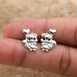 May include: A pair of silver-colored squirrel stud earrings. Each earring features a detailed squirrel design with a heart above its head. The squirrels have black eyes and are facing forward.
