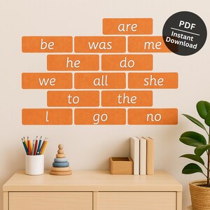 Op de afbeelding: Oranje woordblokken met witte tekst die veelvoorkomende woorden zoals "be", "was", "are" en "the" spellen. Een zwarte cirkel met de tekst "PDF Instant Download" staat rechtsboven. Een houten bureau bevat potloden, een speeltje en boeken.