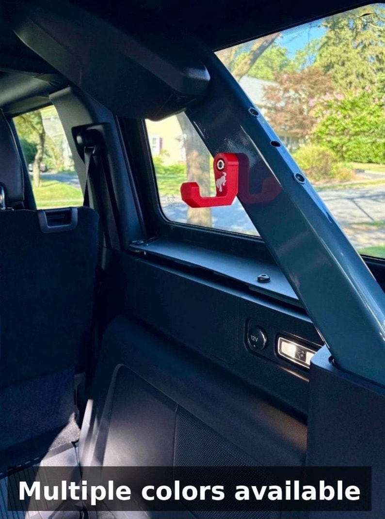 May include: A red metal hook with a horse logo is mounted on a dark gray interior car panel. The hook is near a window with a view of trees and houses. The text "Multiple colors available" is at the bottom.