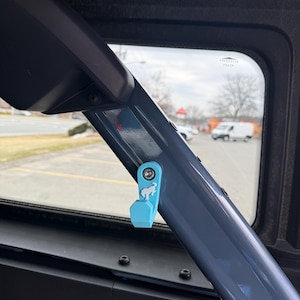 May include: A light blue accessory with a horse logo attached to a dark gray metal bar inside a vehicle. The accessory is mounted near a window, with a view of a street and buildings in the background.