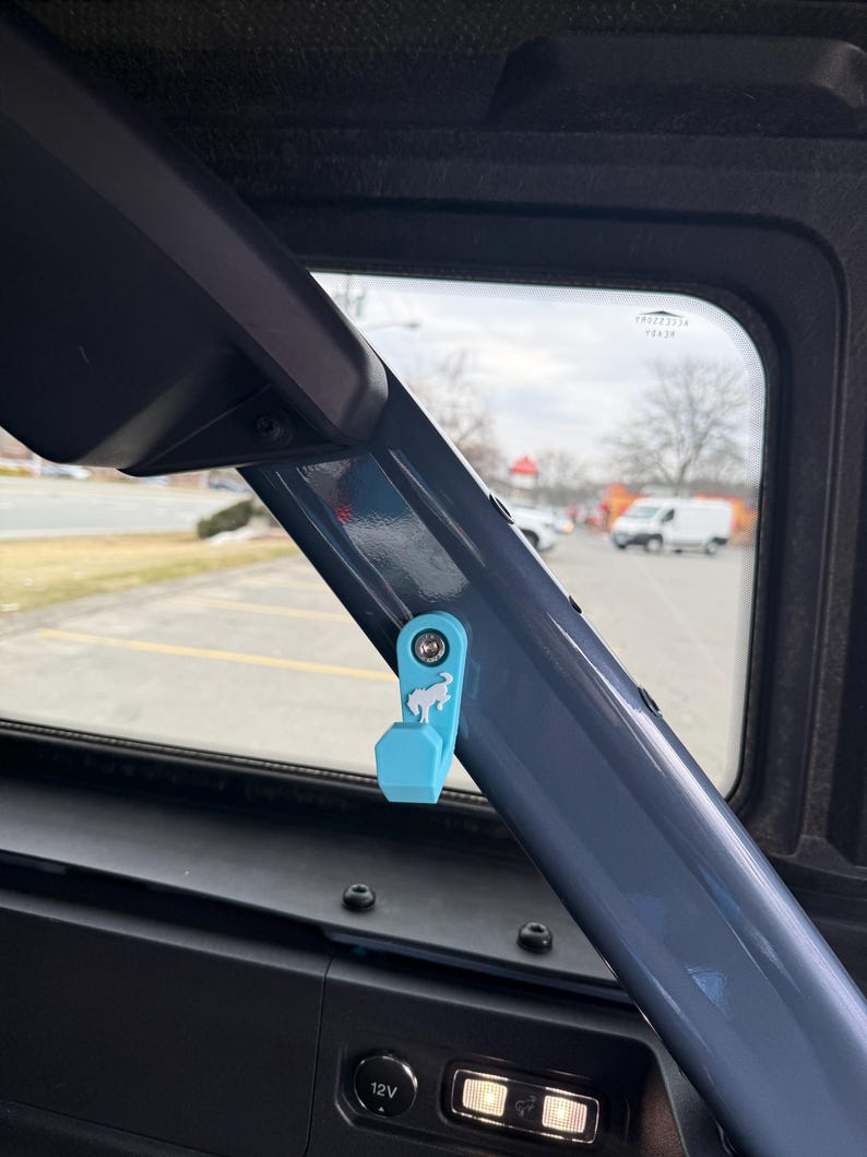 May include: A light blue accessory with a horse logo attached to a dark gray metal bar inside a vehicle. The accessory is mounted near a window, with a view of a street and buildings in the background.