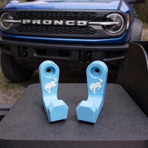 May include: Two light blue, plastic accessories with a white horse logo, designed for a vehicle. The accessories are displayed on a dark surface, with a blue Bronco SUV in the background.