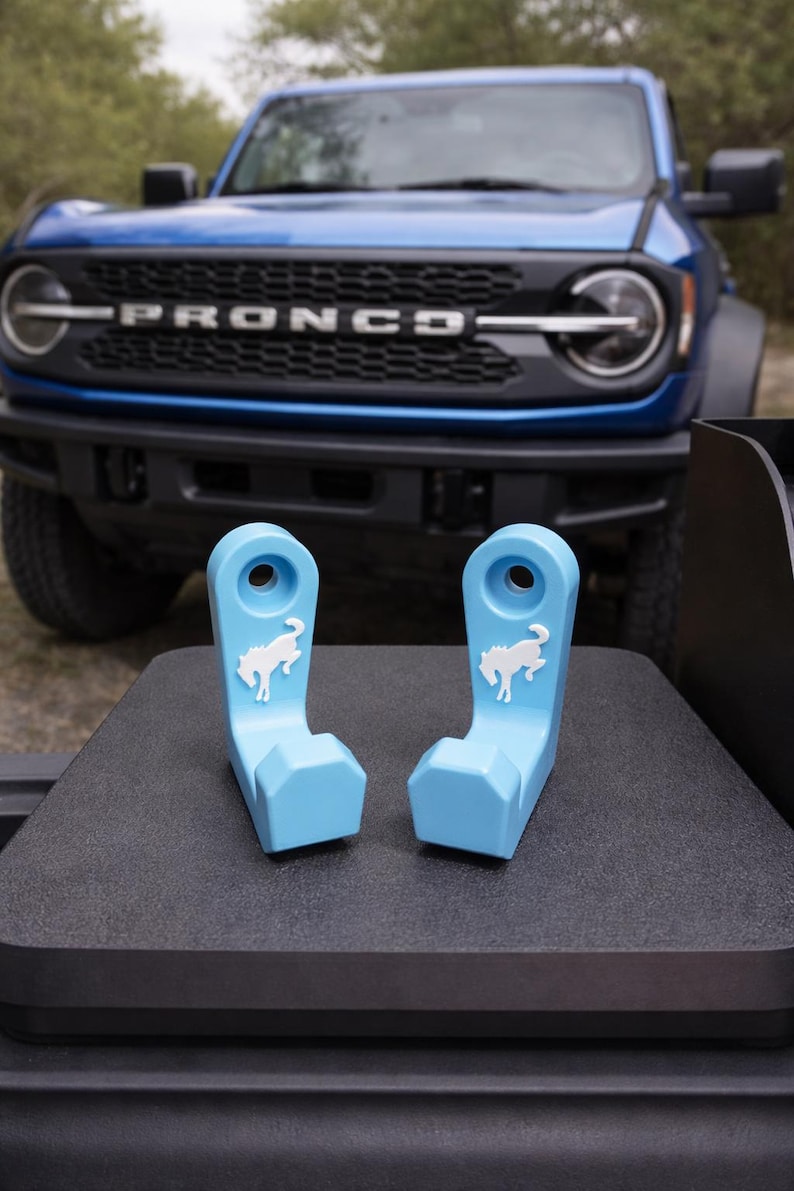 May include: Two light blue, plastic accessories with a white horse logo, designed for a vehicle. The accessories are displayed on a dark surface, with a blue Bronco SUV in the background.