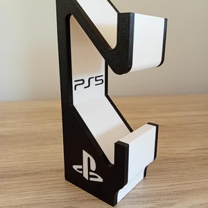 May include: A black and white PS5 console stand. The stand has a curved design with the PS5 logo and PlayStation symbol. The stand is made of a solid material and is designed to hold a gaming console.