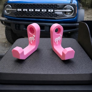 May include: Two bright pink hooks with a white horse logo are displayed on a black surface, with a blue Bronco SUV in the background. The hooks are designed with a curved shape and a hole for hanging.