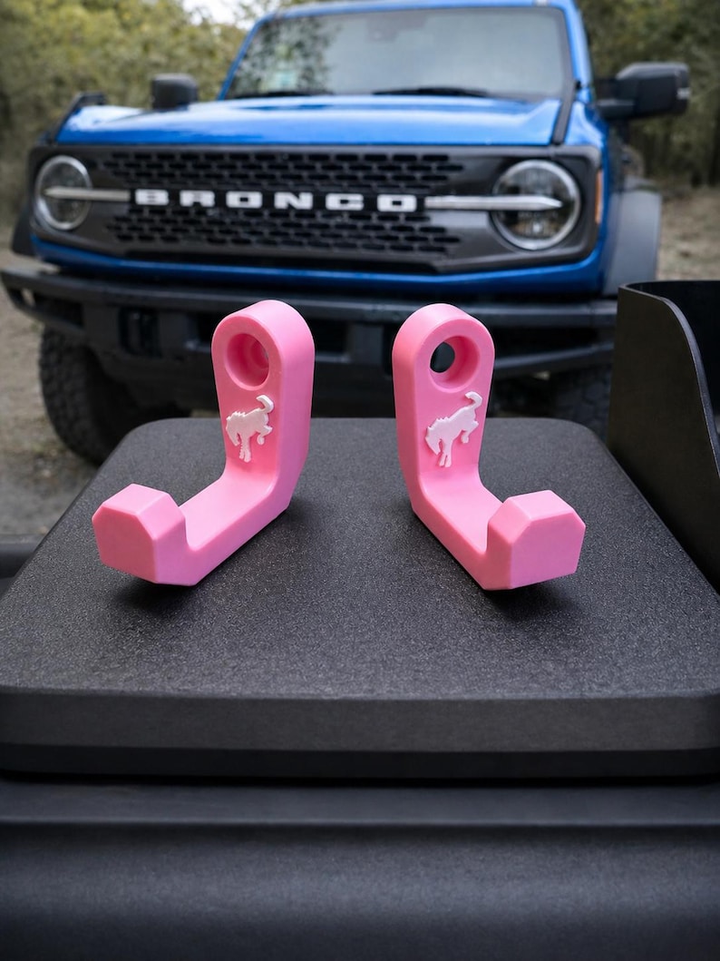 May include: Two bright pink hooks with a white horse logo are displayed on a black surface, with a blue Bronco SUV in the background. The hooks are designed with a curved shape and a hole for hanging.