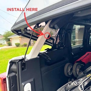 May include: An open car trunk with a white support beam and the text "INSTALL HERE" in red. The trunk contains a stroller and other items. The image is taken outdoors on a sunny day.