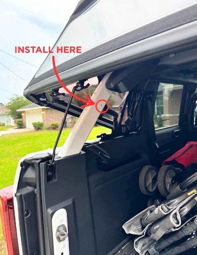 May include: An open car trunk with a white support beam and the text "INSTALL HERE" in red. The trunk contains a stroller and other items. The image is taken outdoors on a sunny day.