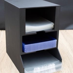 May include: A black, three-tiered storage organizer. The top compartment holds a stack of clear plastic sleeves, the middle holds blue sleeves, and the bottom holds clear sleeves. The organizer is made of a matte black material.