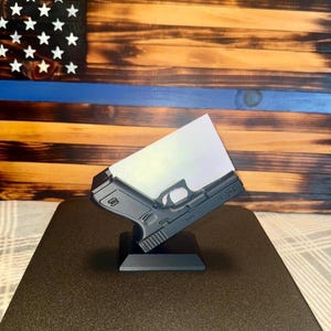 May include: A black gun-shaped display stand holding a white card, set against a wooden American flag with a blue stripe. The stand is on a dark brown surface.