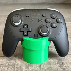 Puede incluir: Un mando Nintendo Switch Pro negro con detalles blancos, apoyado en un soporte en forma de tubo verde brillante. El mando tiene botones X, Y, A y B, y una cruceta. El logotipo de Nintendo Switch es visible.