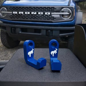 May include: Two blue metal hooks with a white horse logo, likely for a vehicle. The hooks are displayed on a dark surface with a blue SUV in the background. The hooks are designed to attach to a vehicle.
