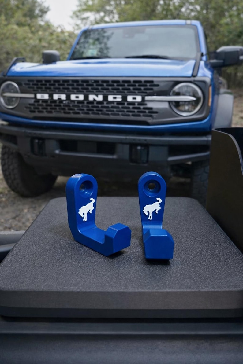 May include: Two blue metal hooks with a white horse logo, likely for a vehicle. The hooks are displayed on a dark surface with a blue SUV in the background. The hooks are designed to attach to a vehicle.