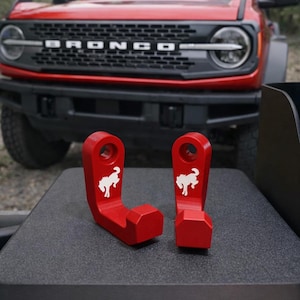 May include: Two red metal hooks with a white horse logo, designed for a vehicle. The hooks are displayed on a dark surface, with a red vehicle in the background.