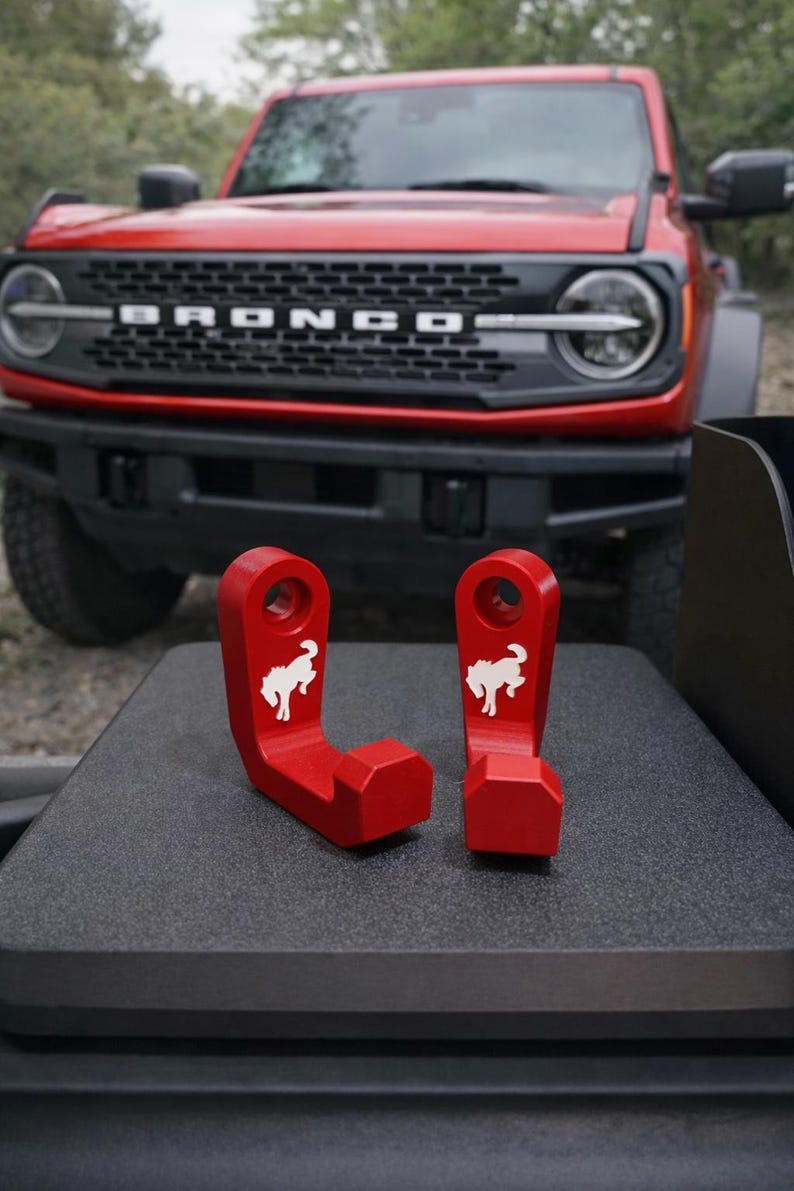 May include: Two red metal hooks with a white horse logo, designed for a vehicle. The hooks are displayed on a dark surface, with a red vehicle in the background.