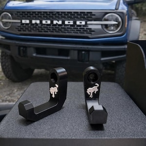 May include: Two black metal hooks with a silver horse logo, displayed in front of a blue Bronco SUV. The hooks are designed with a curved shape and a hexagonal base. The Bronco's front grille is visible.