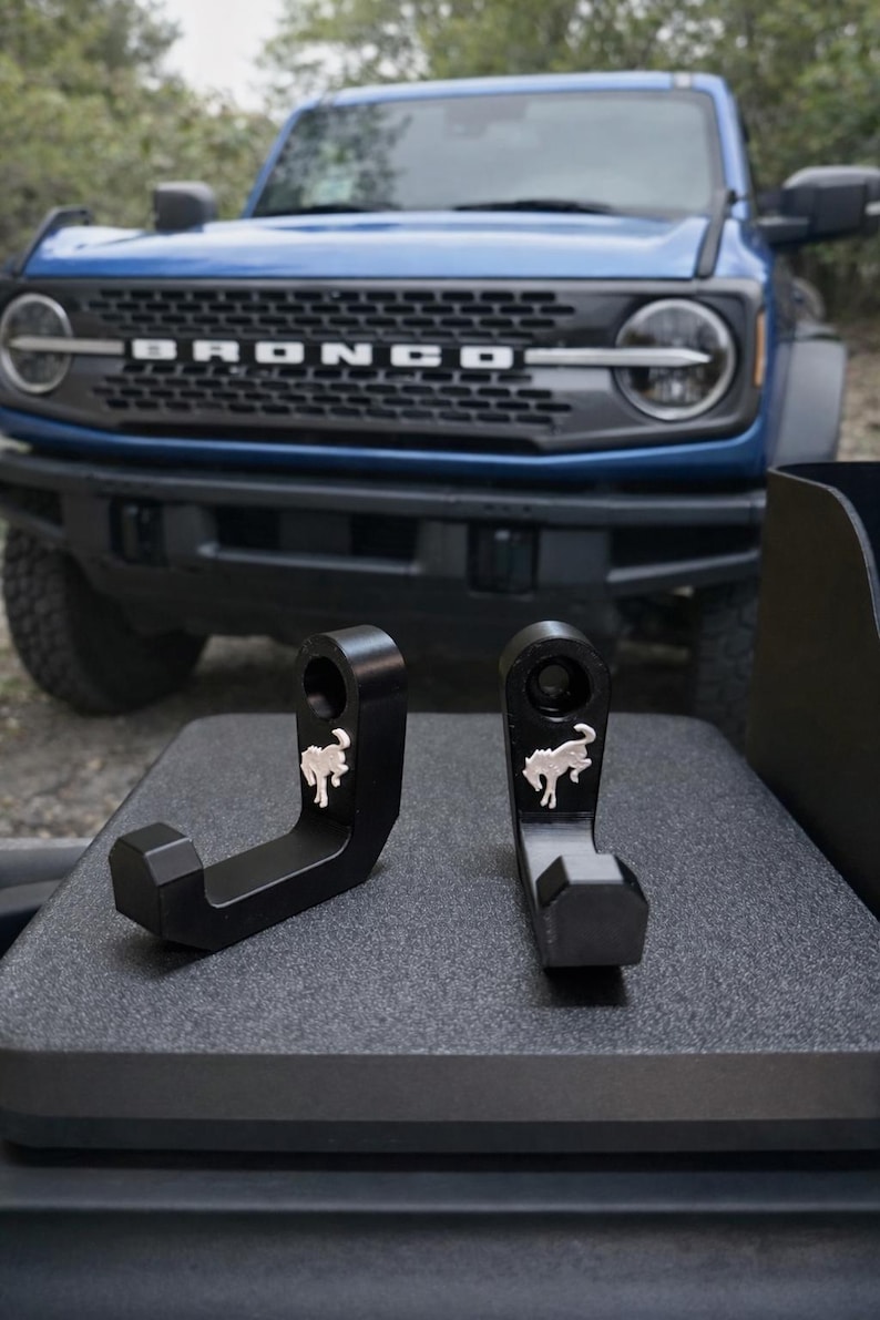 May include: Two black metal hooks with a silver horse logo, displayed in front of a blue Bronco SUV. The hooks are designed with a curved shape and a hexagonal base. The Bronco's front grille is visible.