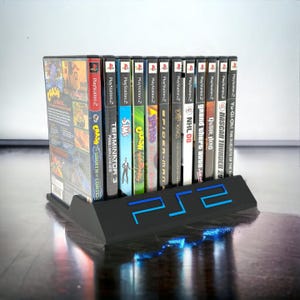 May include: A black plastic display stand featuring the blue PS2 logo, holding a row of twelve PlayStation 2 video game cases. The titles include Crash Bandicoot, Terminator 3, The Sims, Spyro, Spider-Man, Lord of the Rings, NHL 08, Grand Theft Auto, Guitar Hero, NASCAR Thunder, and Yu-Gi-Oh.