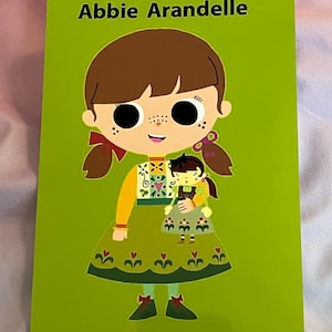 May include: A green book cover with the title "Abbie Arandelle" features a cartoon illustration of a girl holding a doll. The girl has brown hair, freckles, and is wearing a green dress with floral accents. The book's background is a solid green color.