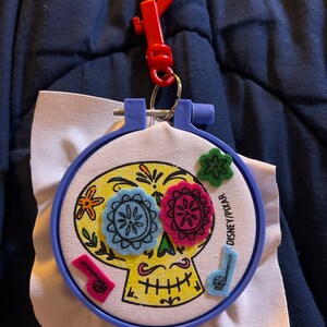 May include: A blue embroidery hoop with a white fabric background featuring a yellow sugar skull with colorful felt accents. The text "DISNEY PIXAR" is printed on the hoop.