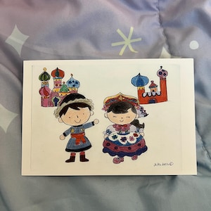May include: A white card featuring a hand-drawn illustration of two cartoon figures in traditional clothing, with colorful buildings in the background. The figures are a boy and a girl. The artist's signature "Aika hello" is in the bottom right corner.
