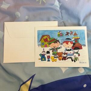 May include: A Christmas-themed greeting card with a colorful illustration of cartoon characters and toys. The card features a drawing of a house, a train, and a sleigh with reindeer. A white envelope is next to the card.