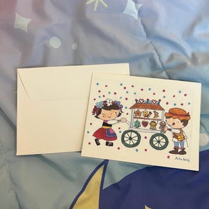 May include: A white envelope and a greeting card with a colorful illustration. The card features two cartoon figures, one pushing a cart with treats. The cart has a sign that says "Love". The card is decorated with polka dots and the artist's signature.