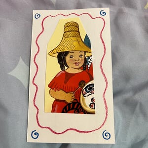 May include: A girl wearing a red dress and a large, woven hat is holding a drum with a colourful design. The image is framed with a pink and blue border.